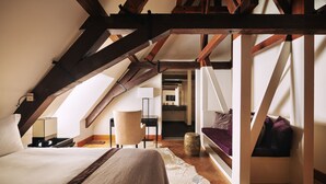 A bedroom with a sloped ceiling, wooden beams, a bed, a nightstand, a lamp, a chair, and a view into the bathroom. - Hotel Prinsenhof (Groningen)