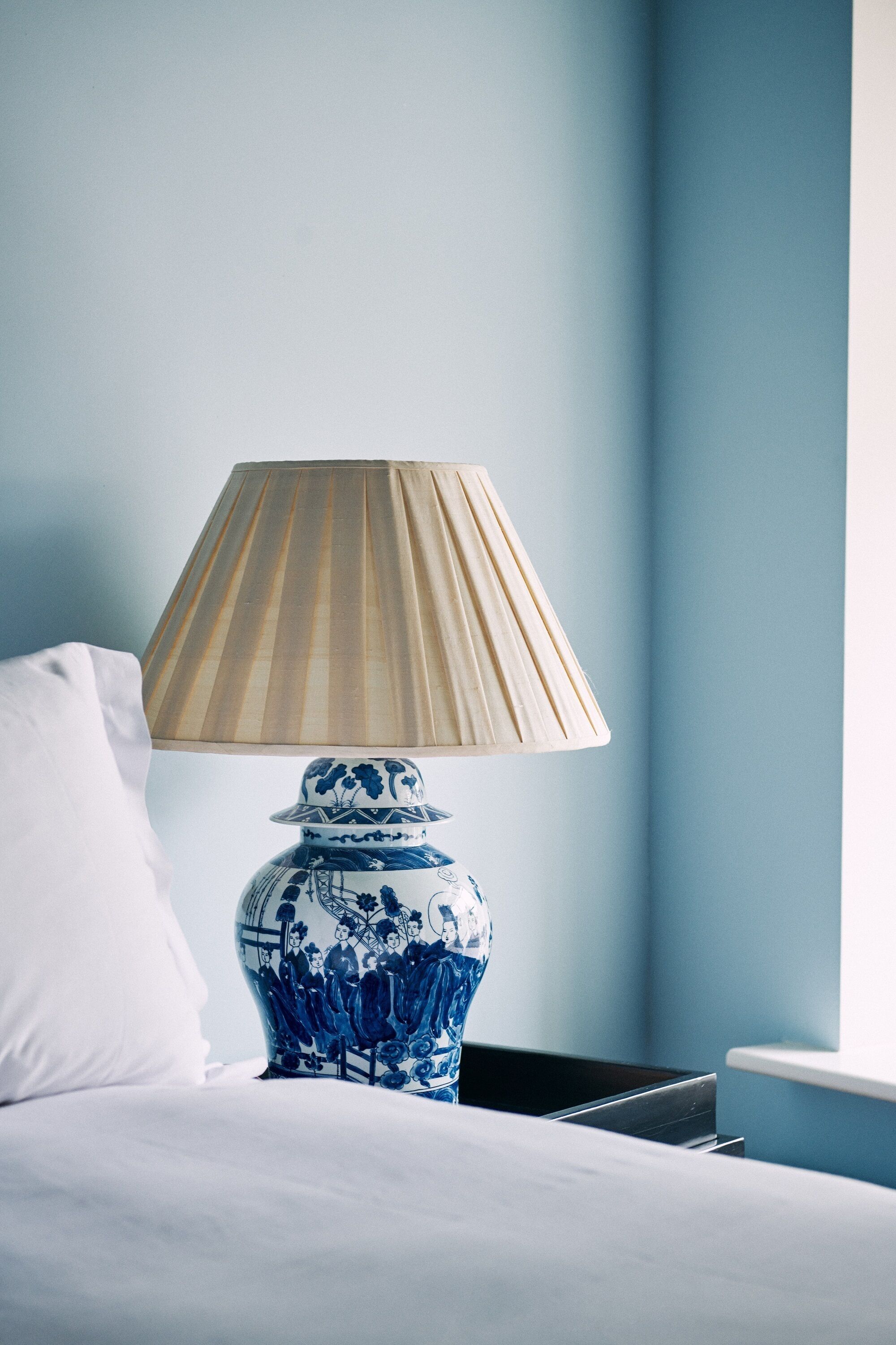 Una cama con un almohadón blanco, una lámpara de cerámica azul y blanca con pantalla color crema, y una pared azul.