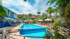 Una piscina con sillones de descanso y sombrillas, rodeada de vegetación y edificios.