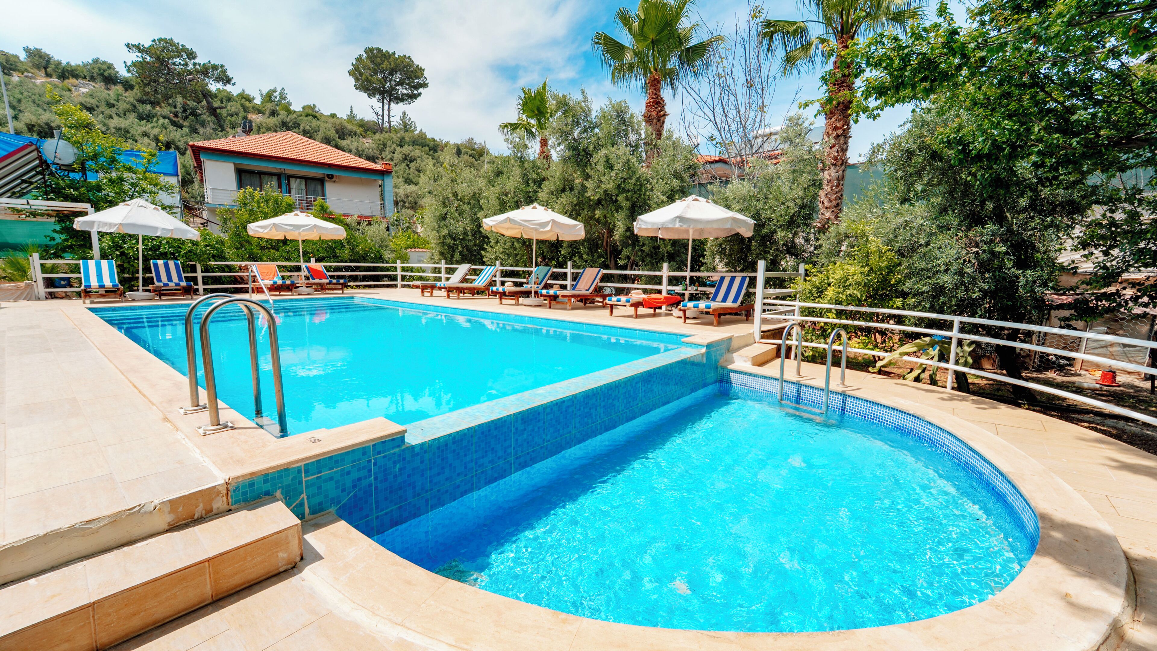 Una piscina con sillas de descanso y sombrillas, rodeada de árboles y un edificio al fondo.