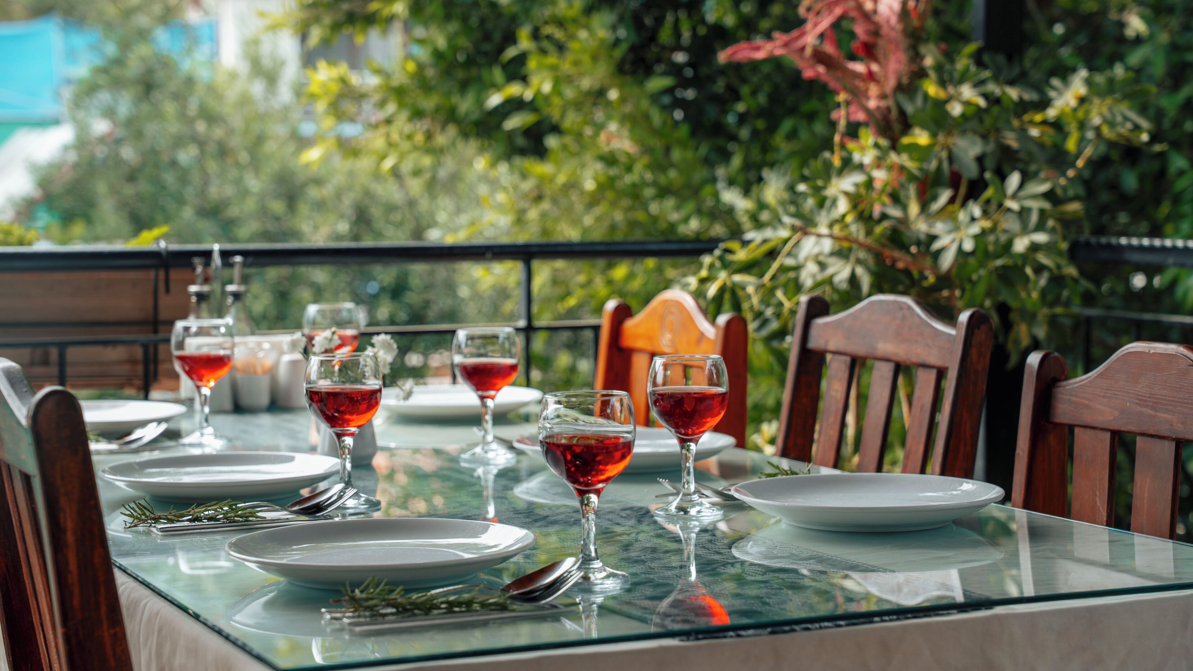 Une table en verre, ornée de verres à vin et d’assiettes, entourée de chaises en bois, installée à l’extérieur avec une végétation luxuriante en arrière-plan.