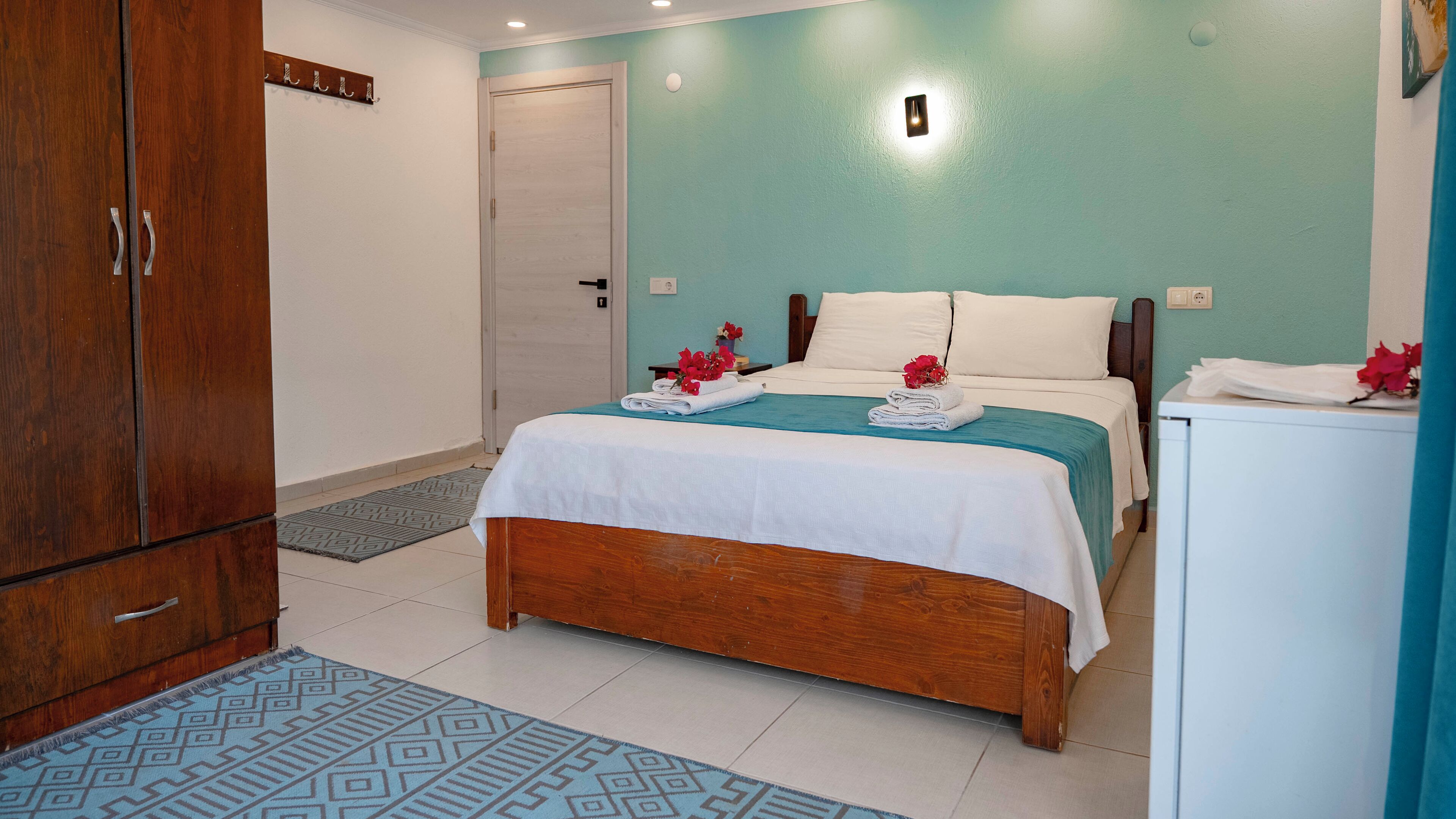 A bedroom with a bed, a wooden wardrobe, a small table with a vase, and a wall-mounted light.