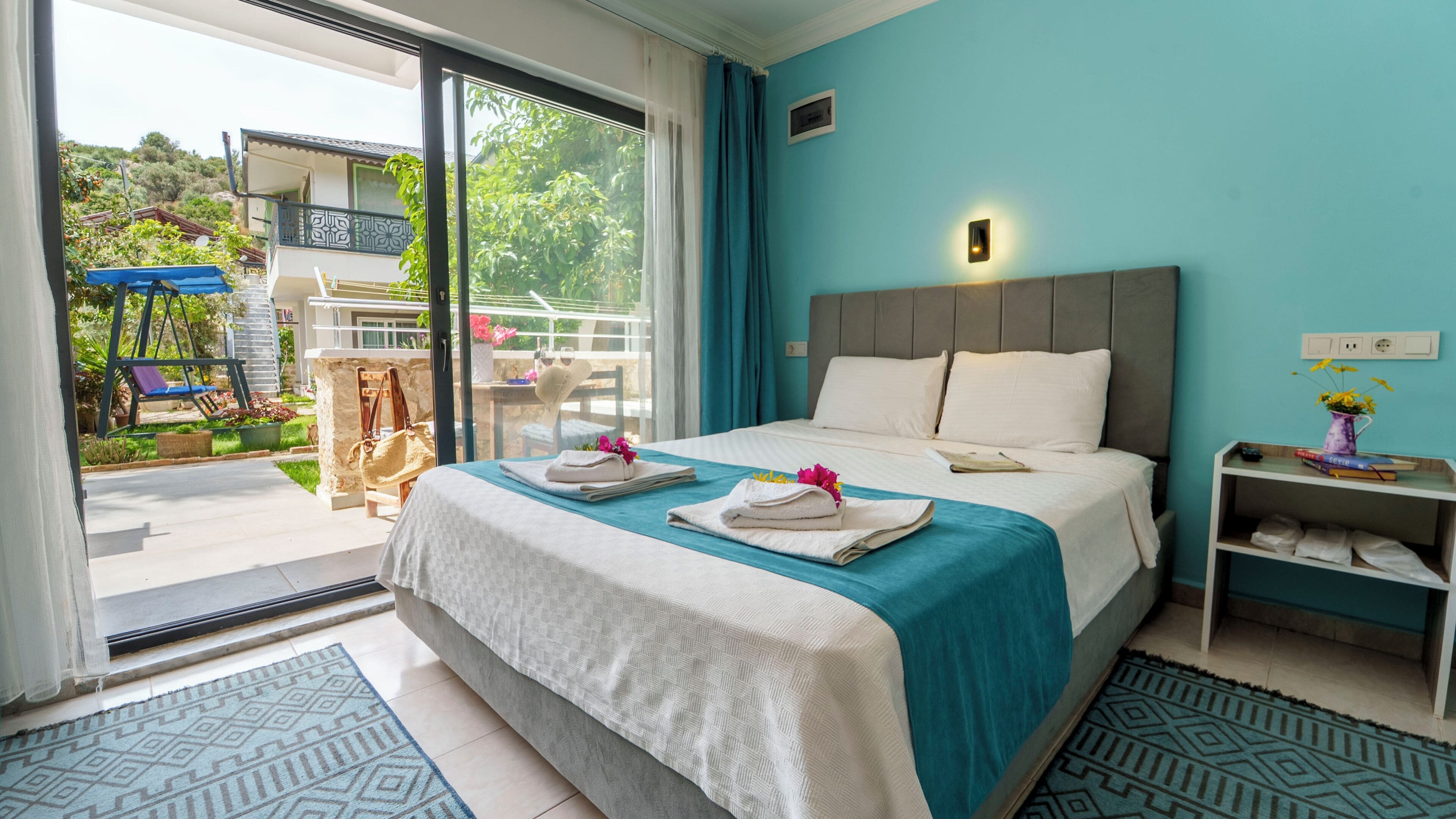 A hotel room with a bed, a small bedside table, a view of the outside, and a balcony with outdoor furniture.