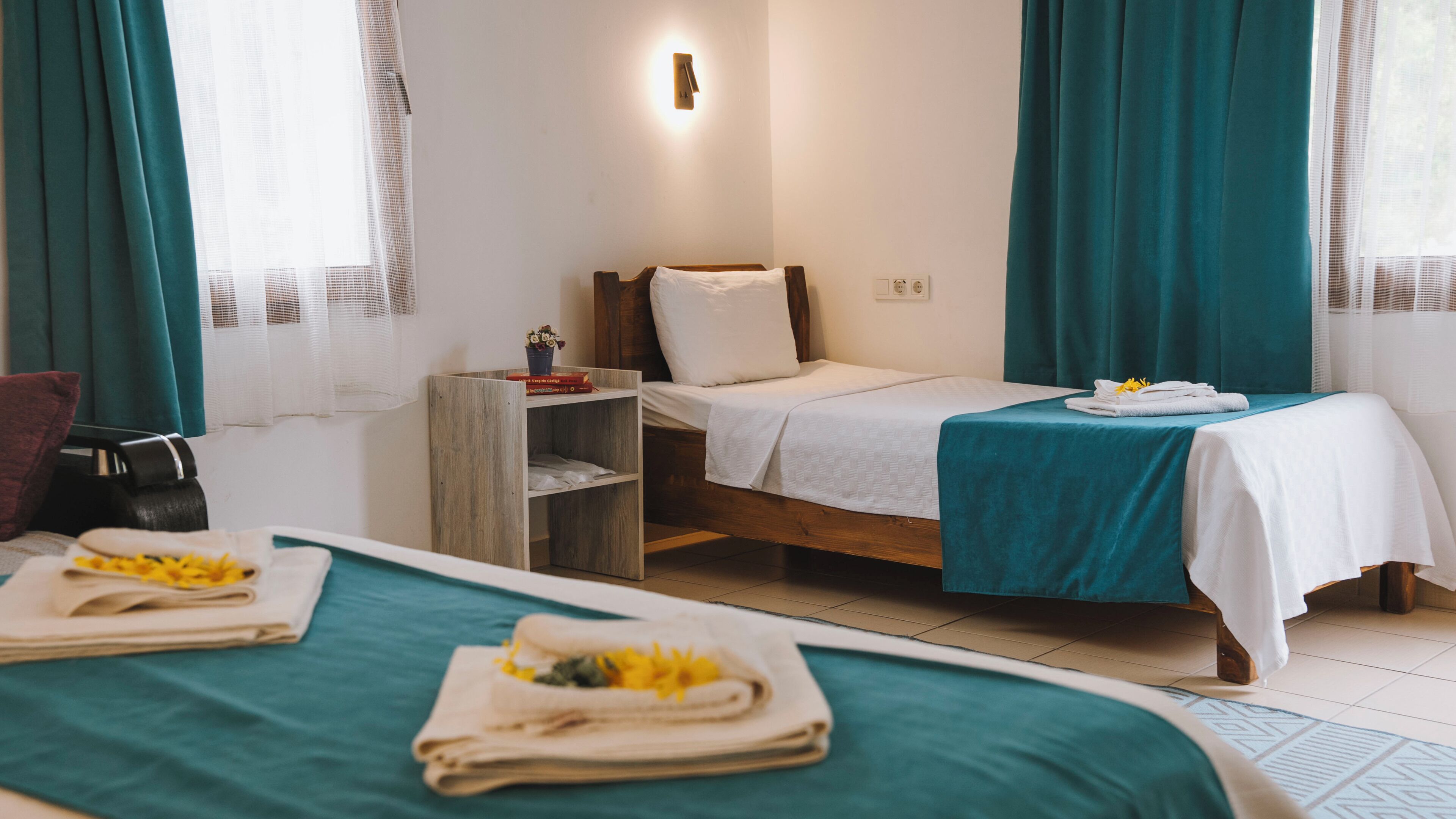 Camera d'albergo con due letti, un comodino in legno, una lampada a parete e una finestra con tende.