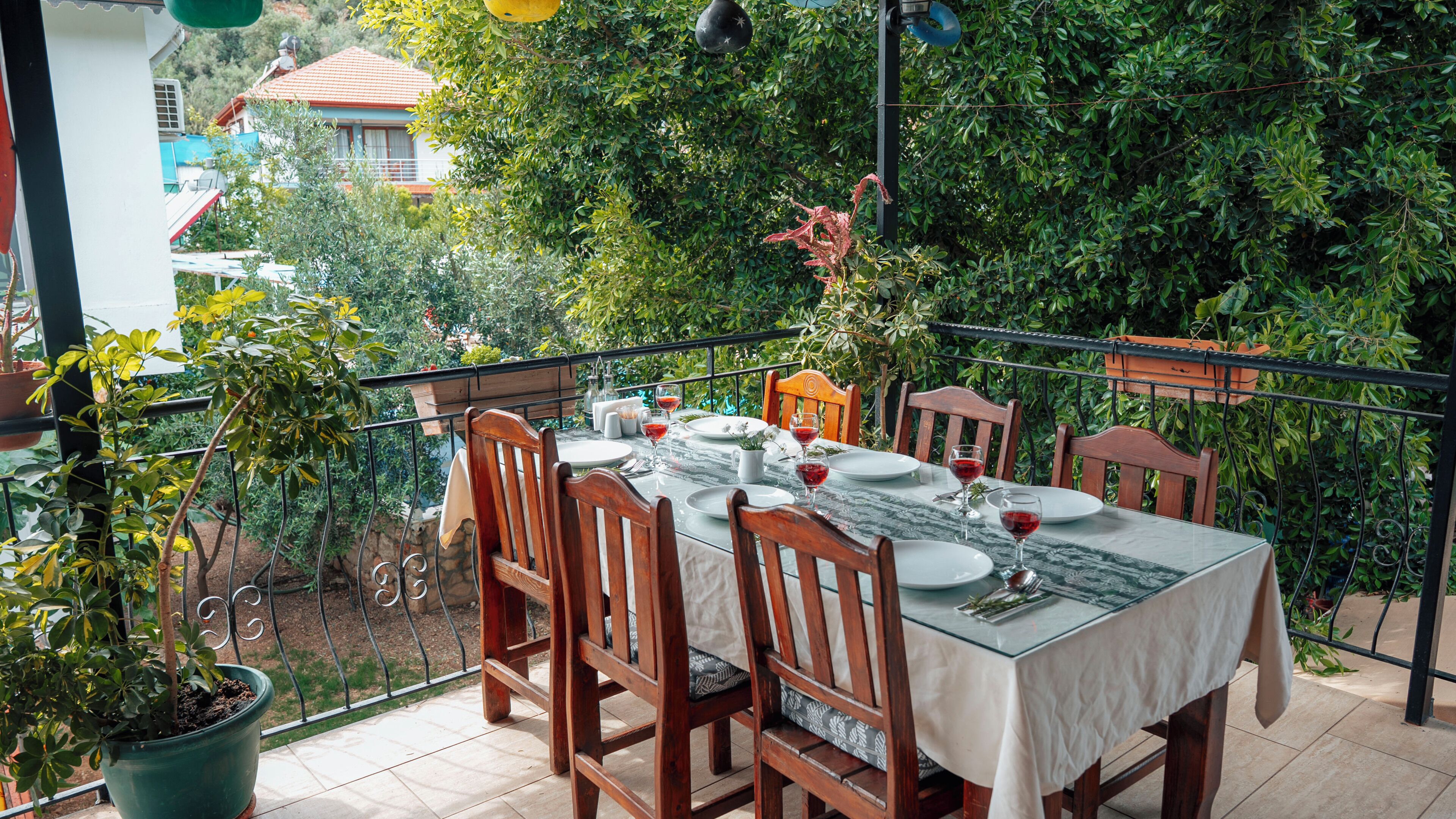 Un área de comedor al aire libre con una mesa dispuesta para cuatro personas, rodeada de sillas de madera, plantas en macetas y vistas a una zona residencial.