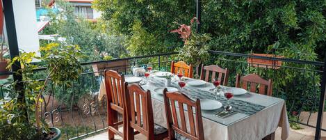 Un área de comedor al aire libre con una mesa dispuesta para cuatro personas, rodeada de sillas de madera, plantas en macetas y vistas a una zona residencial.