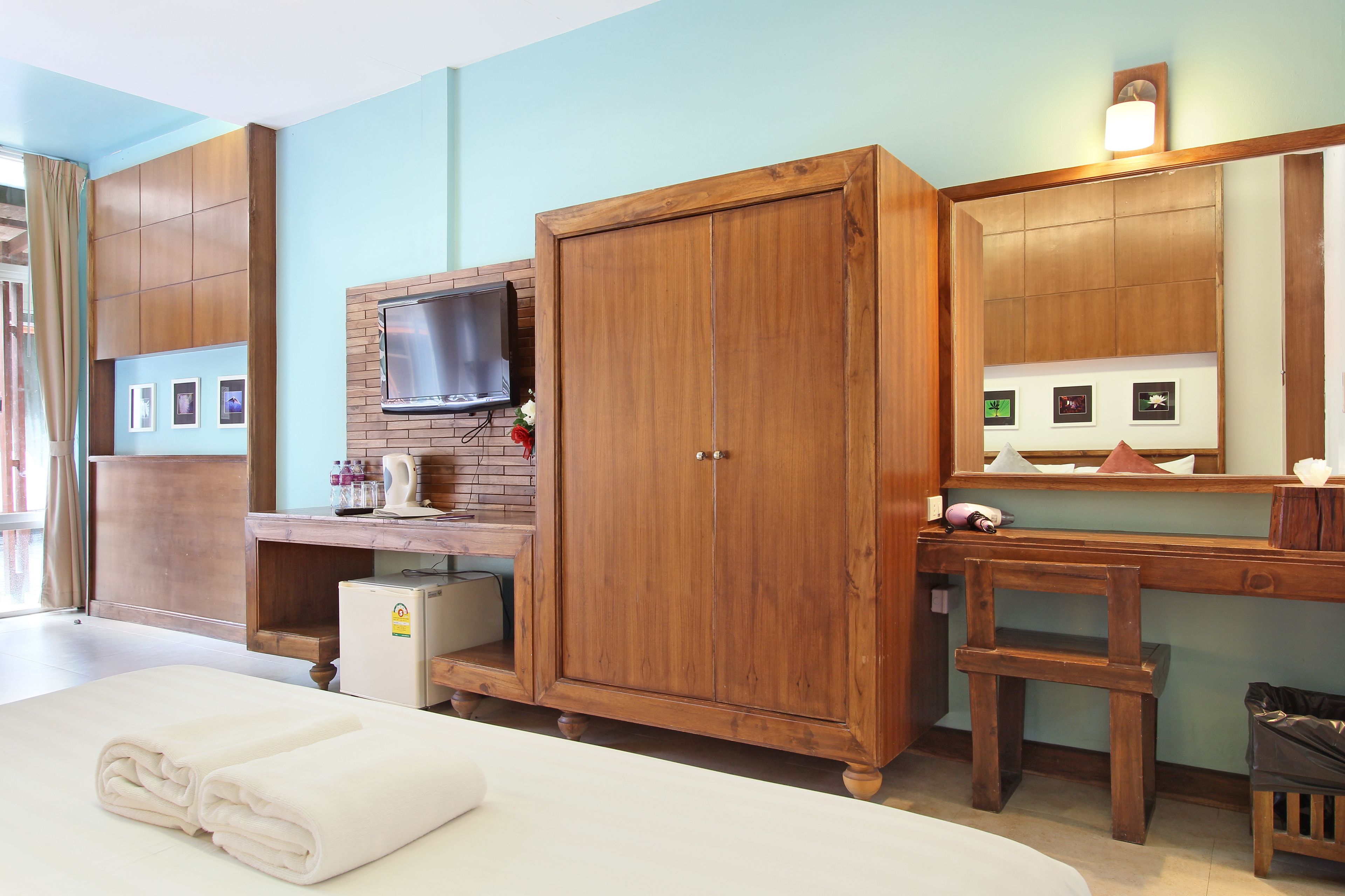 A hotel room with a wooden wardrobe, a TV mounted on the wall, a small desk with a computer, and a bed with neatly folded towels.