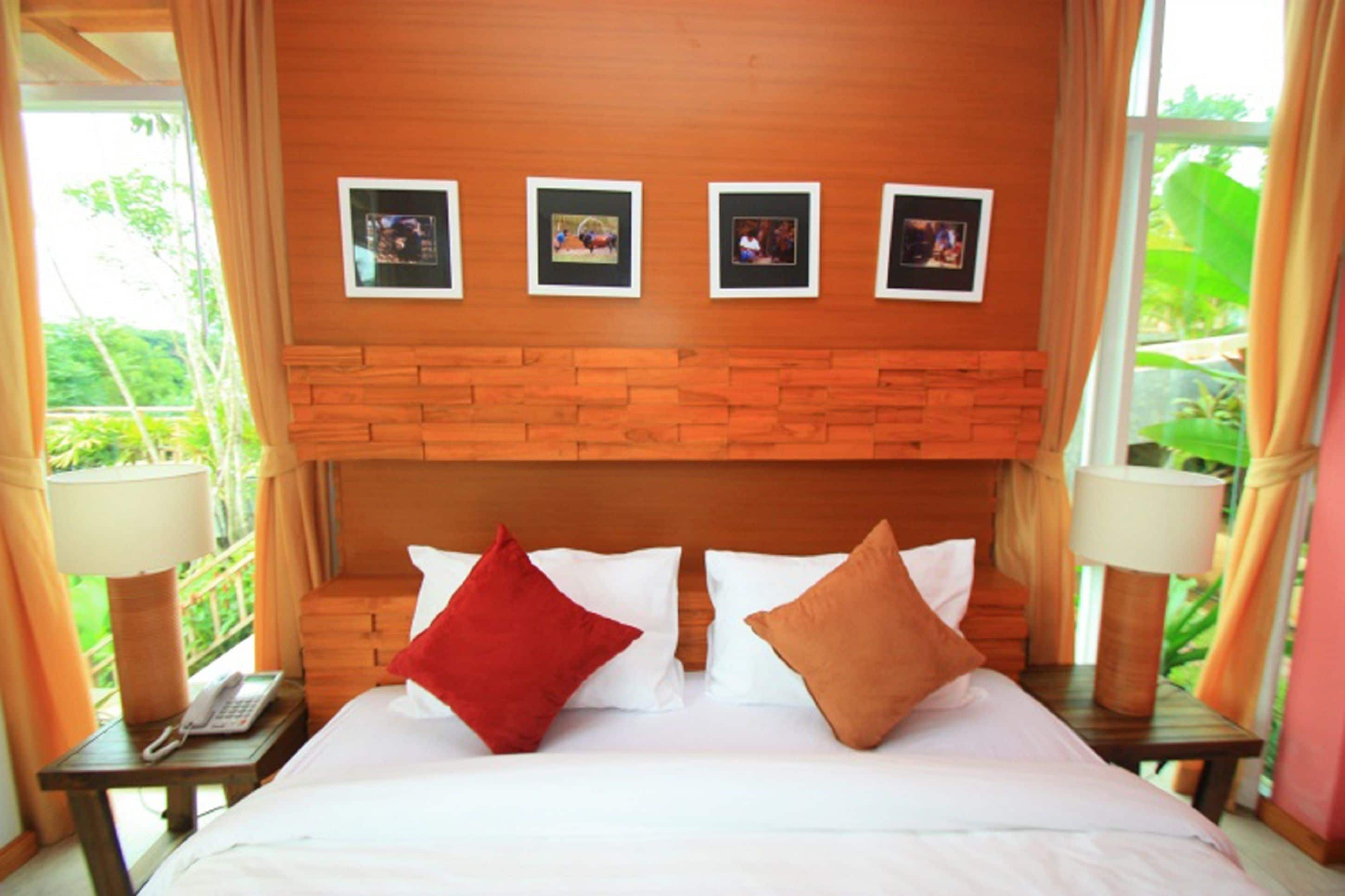 A hotel room with a wooden headboard, a bed with white and red pillows, a nightstand with a lamp, and a window with curtains.