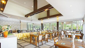 A spacious dining area with multiple tables and chairs, large windows offering a view of greenery outside.