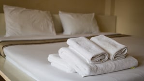 A neatly made bed with white linens and pillows, a folded towel on the bed, and a plain wall in the background. - Triple Palms House (Chiang Mai)