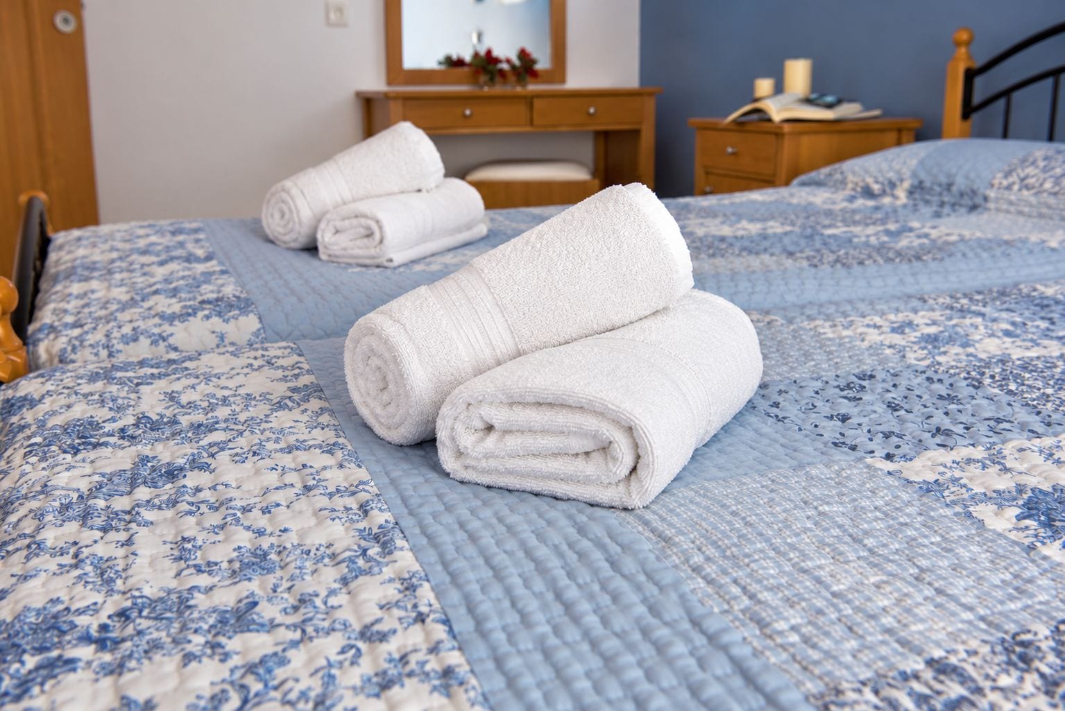 Un lit bien fait, avec des serviettes blanches pliées en rouleaux, une couvre-lit bleue et blanche à motifs, et une table de chevet en bois sur laquelle se trouvent un livre et une lampe.