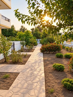 A landscaped garden with a paved walkway, various shrubs, and a building with a balcony. - Nephele Apartments (Rhodes)