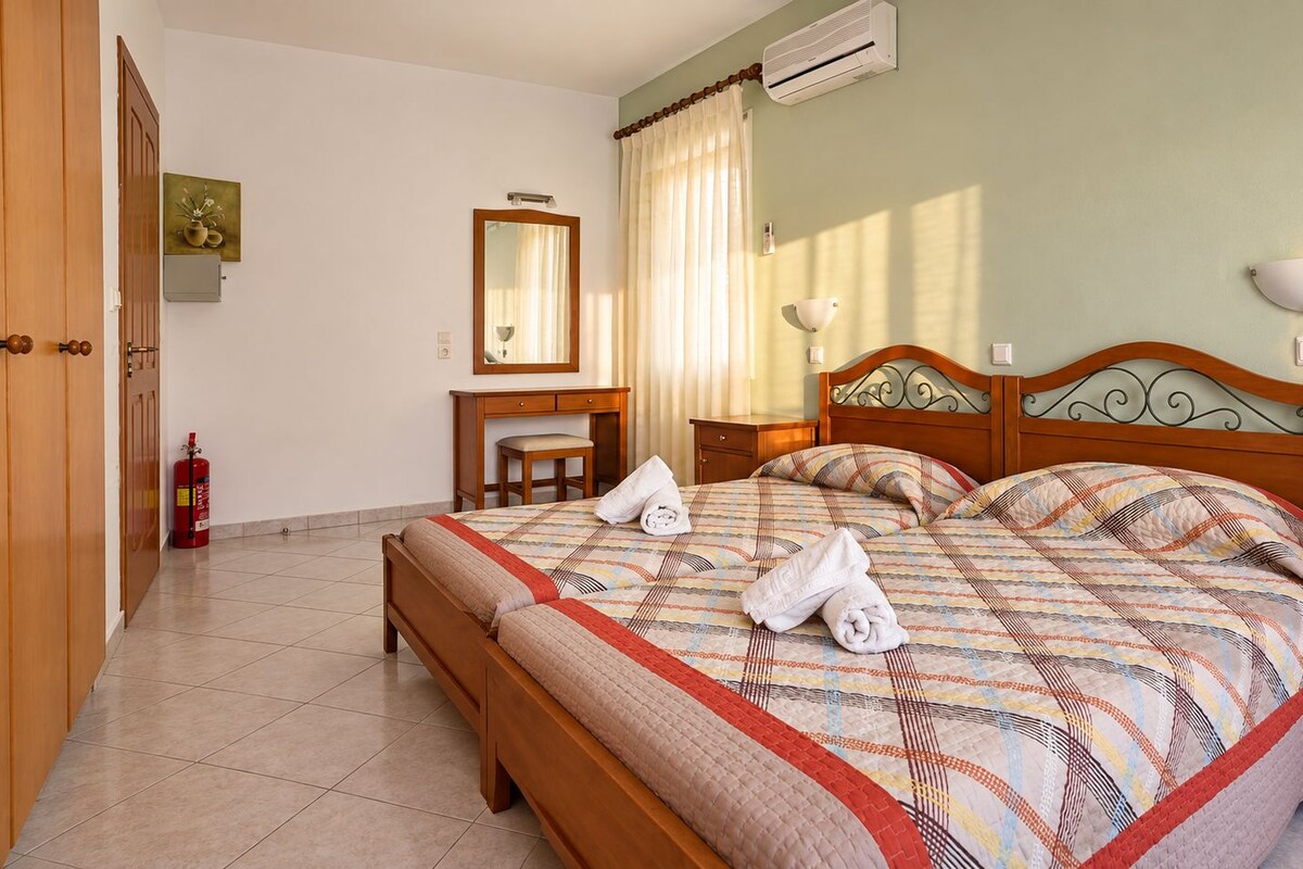 Una habitación de hotel con cama, mesitas de noche, escritorio, silla, extintor y ventana con cortinas.