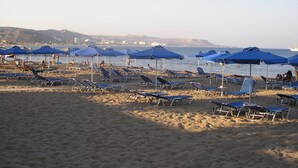 A beach with rows of sun loungers and blue umbrellas, a clear sky, and a distant view of hills and buildings. - Nephele Apartments (Rhodes)