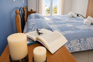 A bedroom with a bed, a bedside table with a book and glasses, two candles, and a view of the outdoors. - Nephele Apartments (Rhodes)