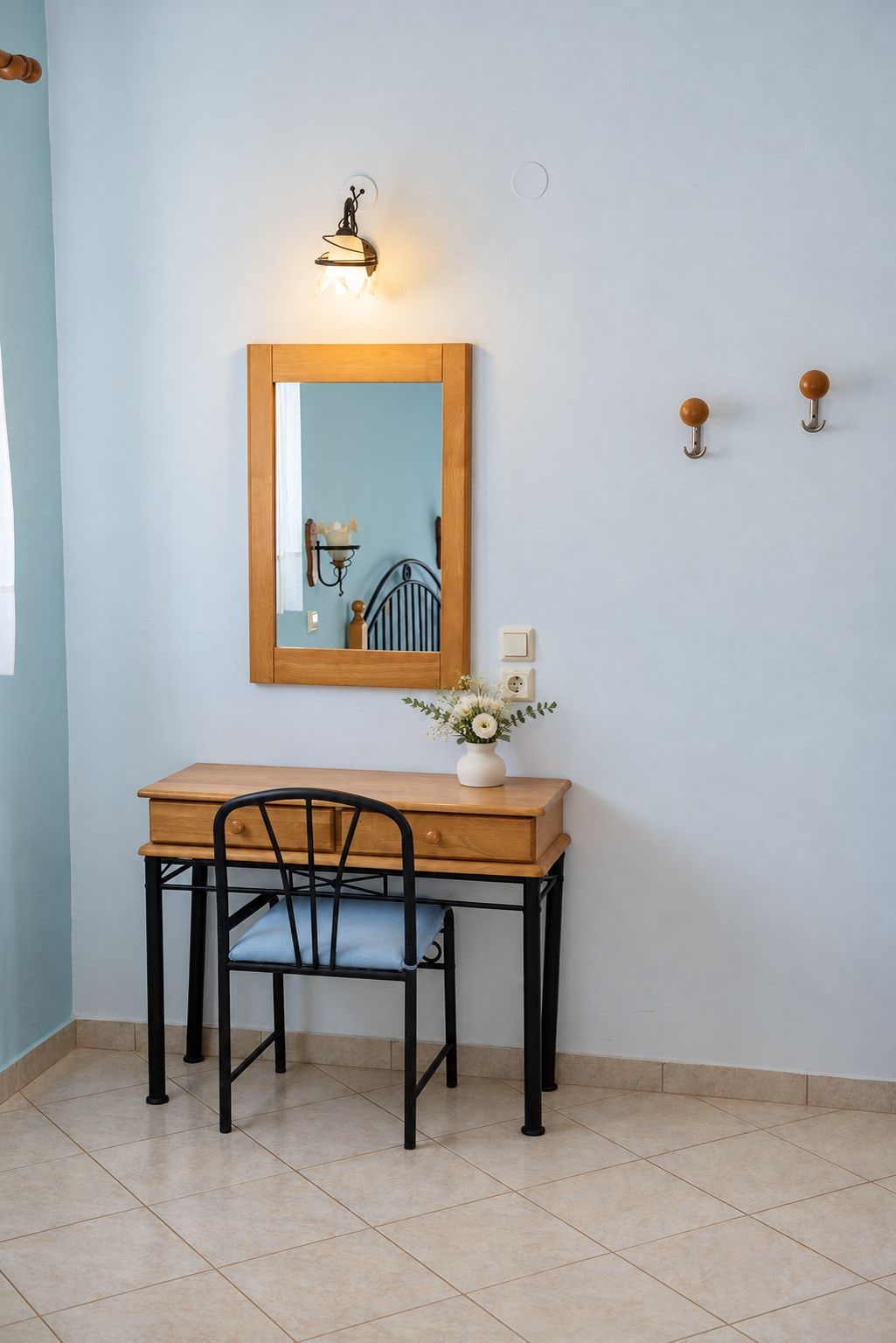 A small room with a wooden desk, a chair, a wall-mounted mirror, a wall-mounted light, and a small vase with flowers.