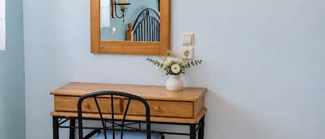 A small room with a wooden desk, a chair, a wall-mounted mirror, a wall-mounted light, and a small vase with flowers.