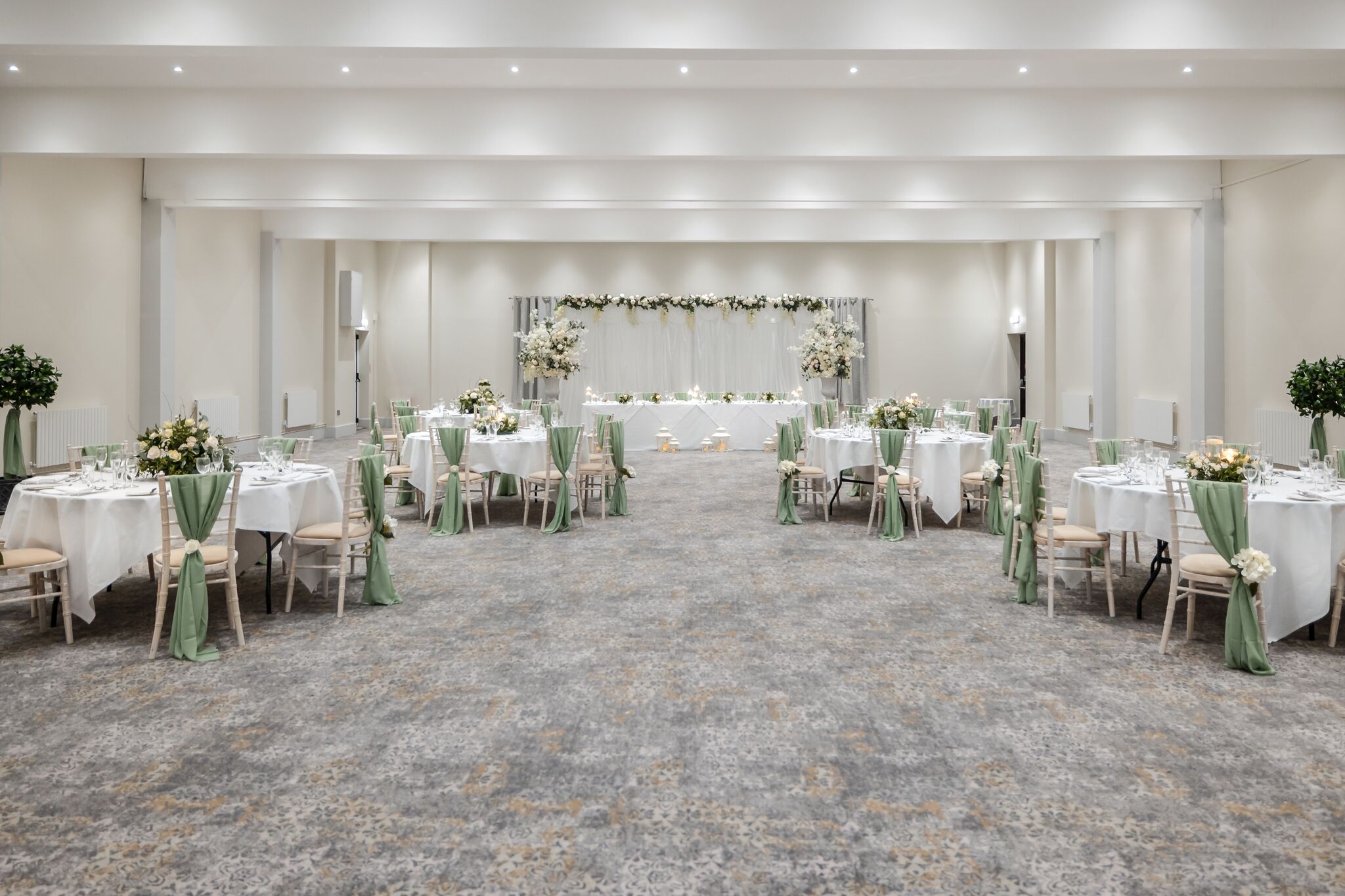 Een feestzaal met ronde tafels, groene stoelhoezen en bloemrijke decoraties.