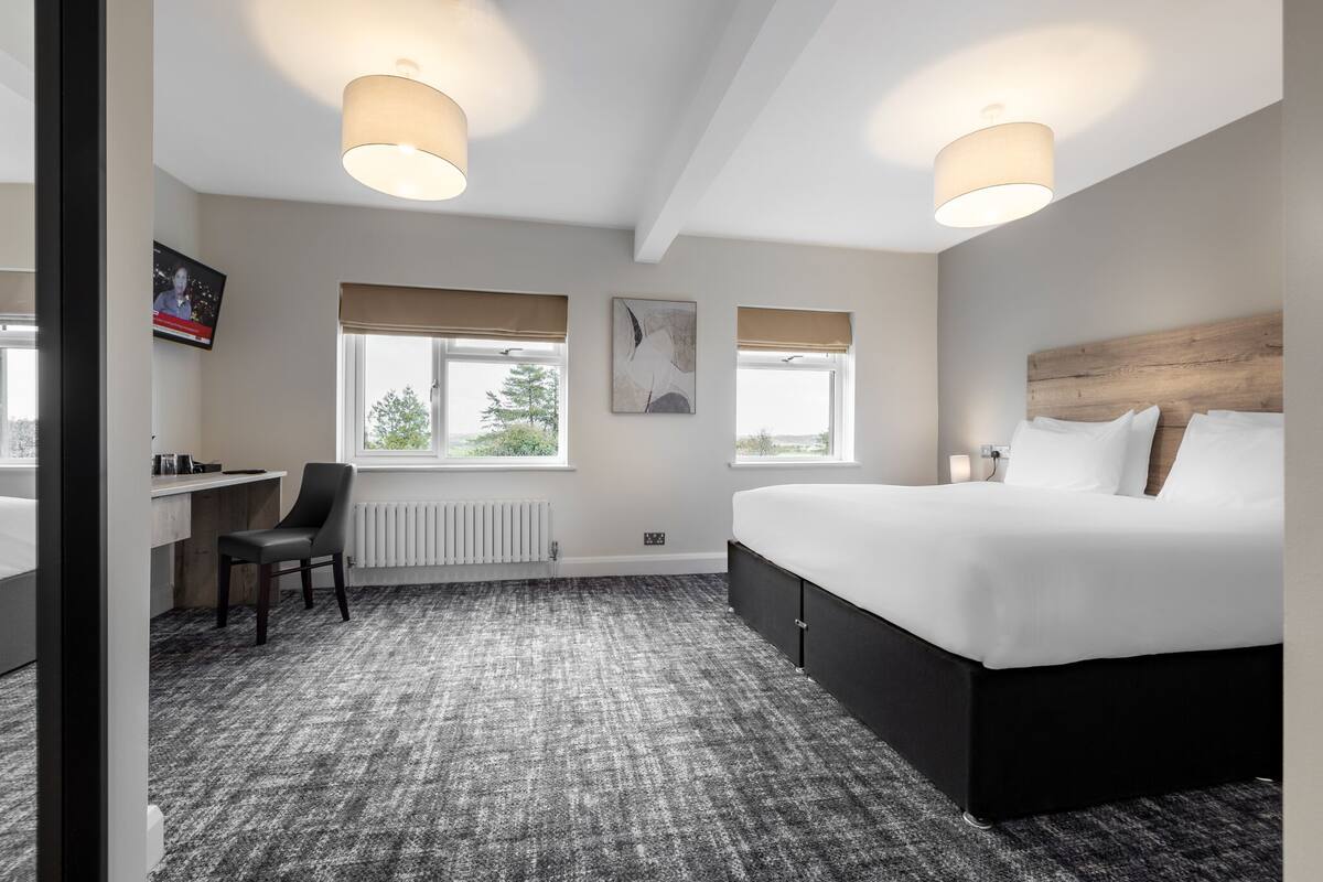 Quarto de hotel moderno com uma cama grande, uma escrivaninha e duas janelas com cortinas bege.
