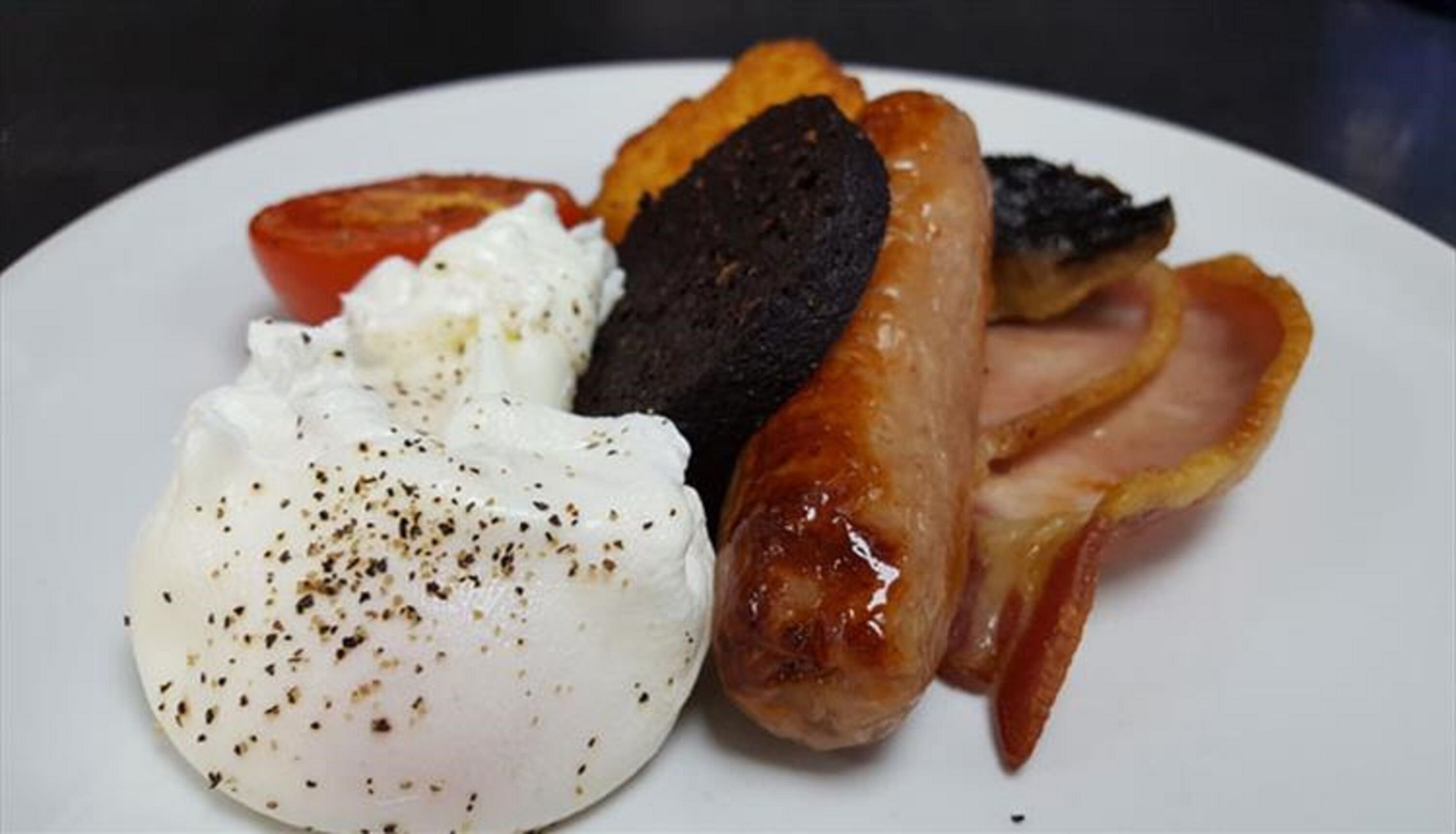 A plate with a poached egg, a grilled sausage, a slice of tomato, and a piece of bacon.