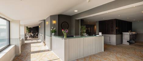 A spacious hotel lobby with a reception desk, a clock, and a dining area in the background.