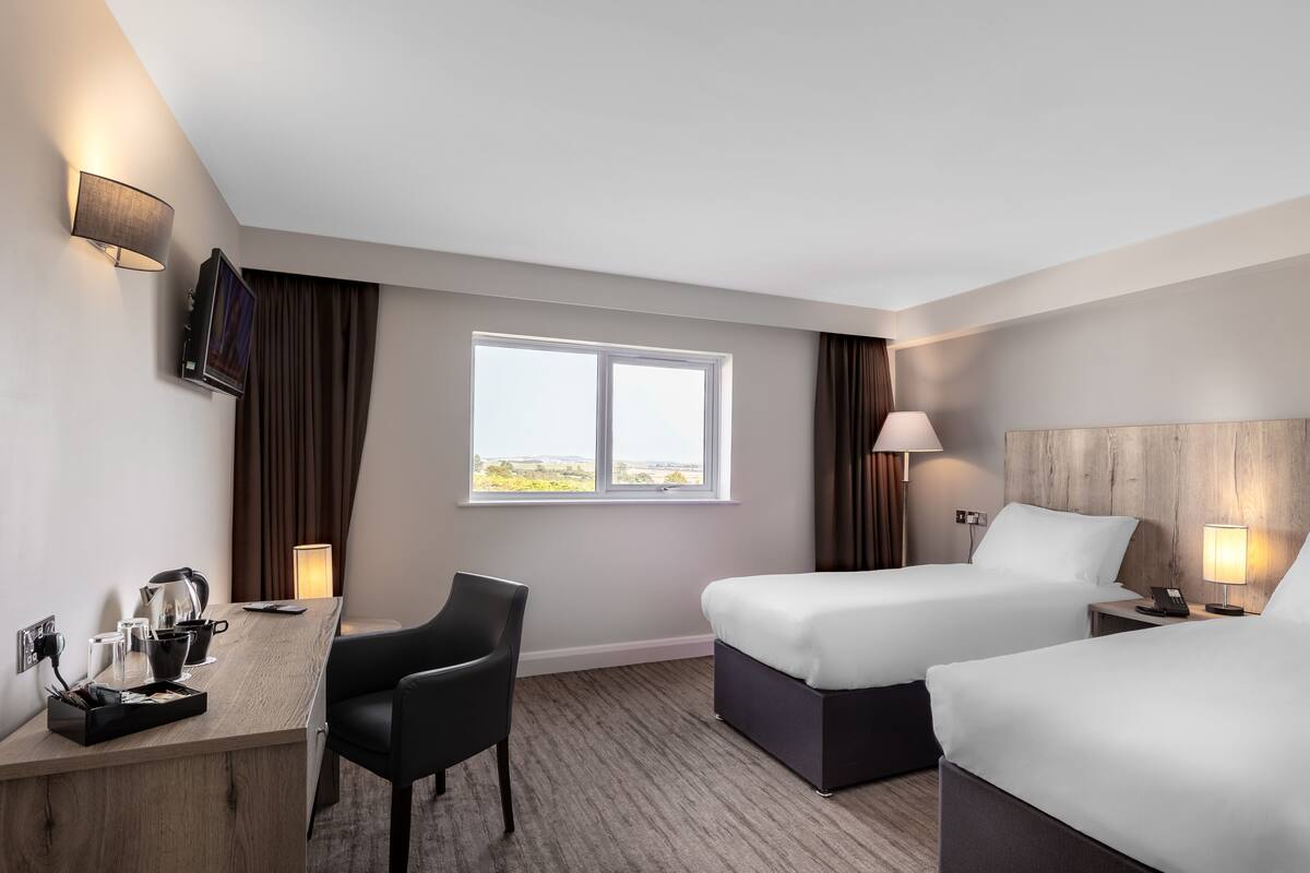 Quarto de hotel com duas camas, uma escrivaninha, uma cadeira, uma televisão e janela com vista.