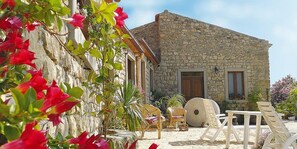 A stone building with a rustic exterior, outdoor seating area with chairs and a table, and a garden with red flowers. - San Noto Turismo Rurale (Ficarra)