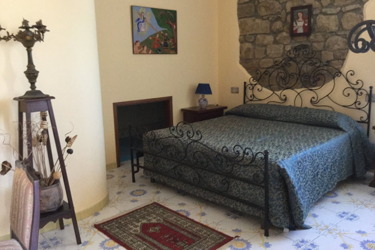 Une chambre avec un mur en pierre, un lit recouvert d’une couvre-lit à motifs, une table de chevet avec une lampe, une plante décorative et un tableau accroché au mur.