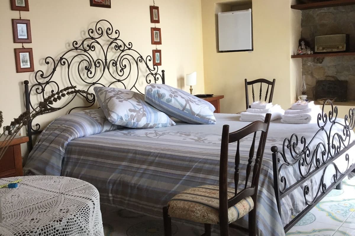 Une chambre à coucher avec un lit, une chaise, une petite table recouverte d’une nappe au crochet et un mur orné de tableaux encadrés.