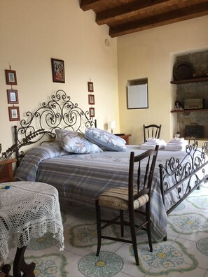 A bedroom with a bed, a chair, a small table with a crocheted tablecloth, and a wall with framed pictures.