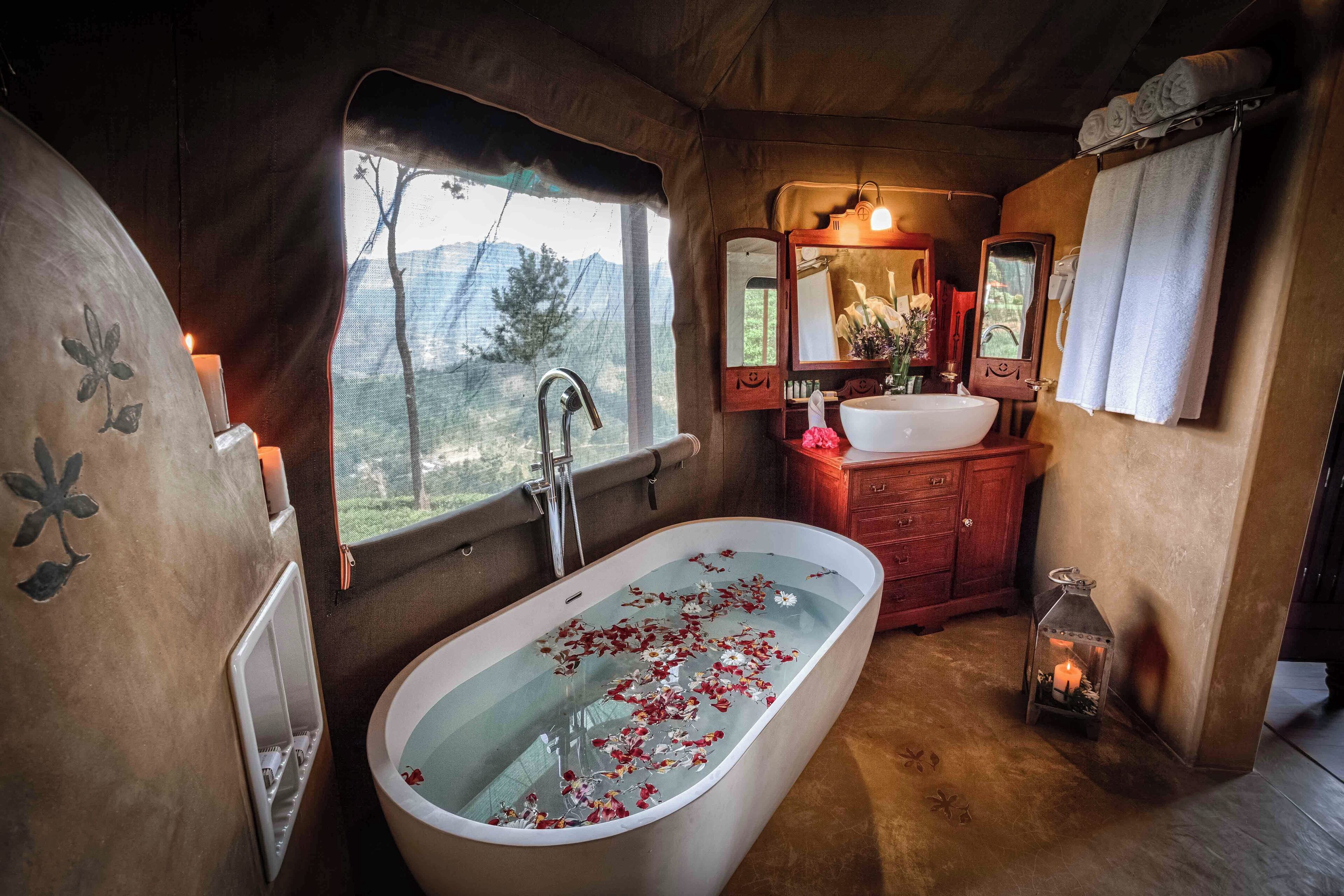A bathroom with a freestanding bathtub filled with rose petals, a window with a view of trees, a wooden vanity with a mirror, and a candle on the floor.