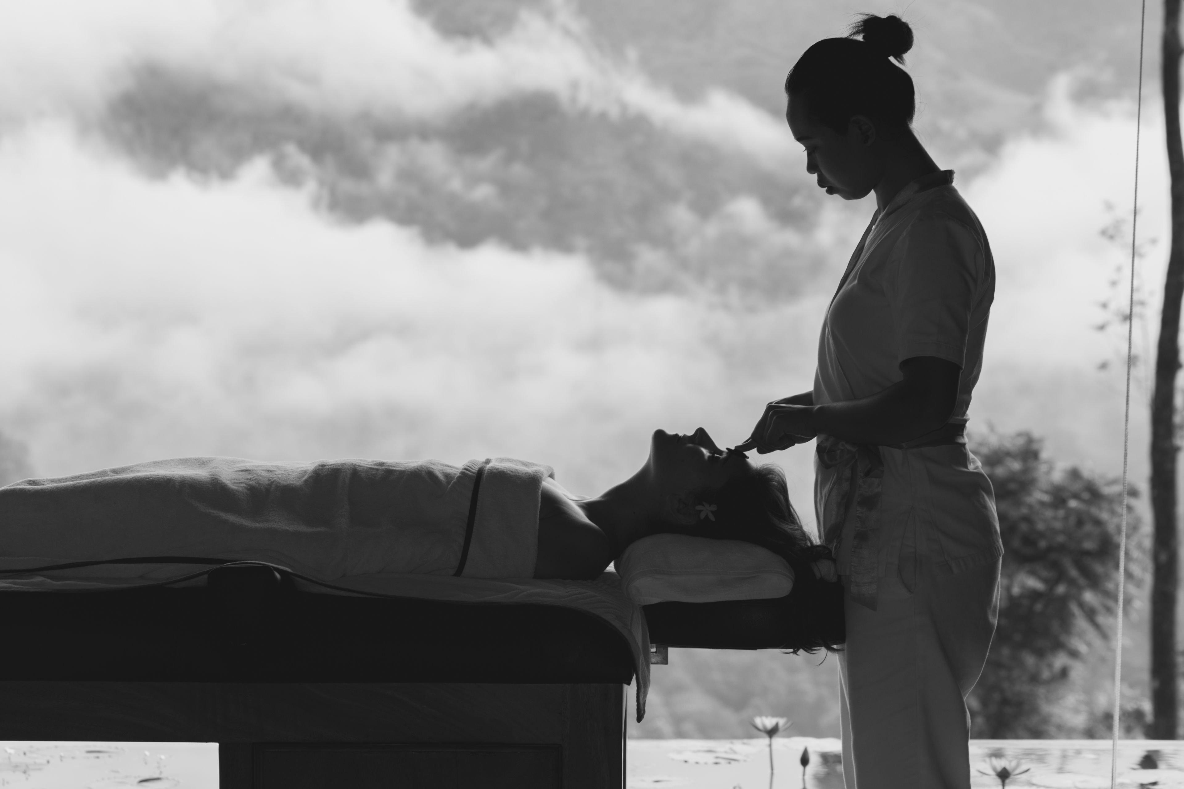 A person attending to another individual on a bed in a room with a view of the sky.