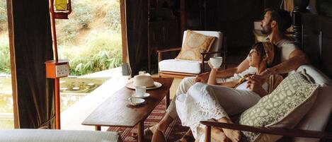 A cozy room with a bed, a table, and chairs. There is a window with curtains, a lamp, and a view of greenery outside.