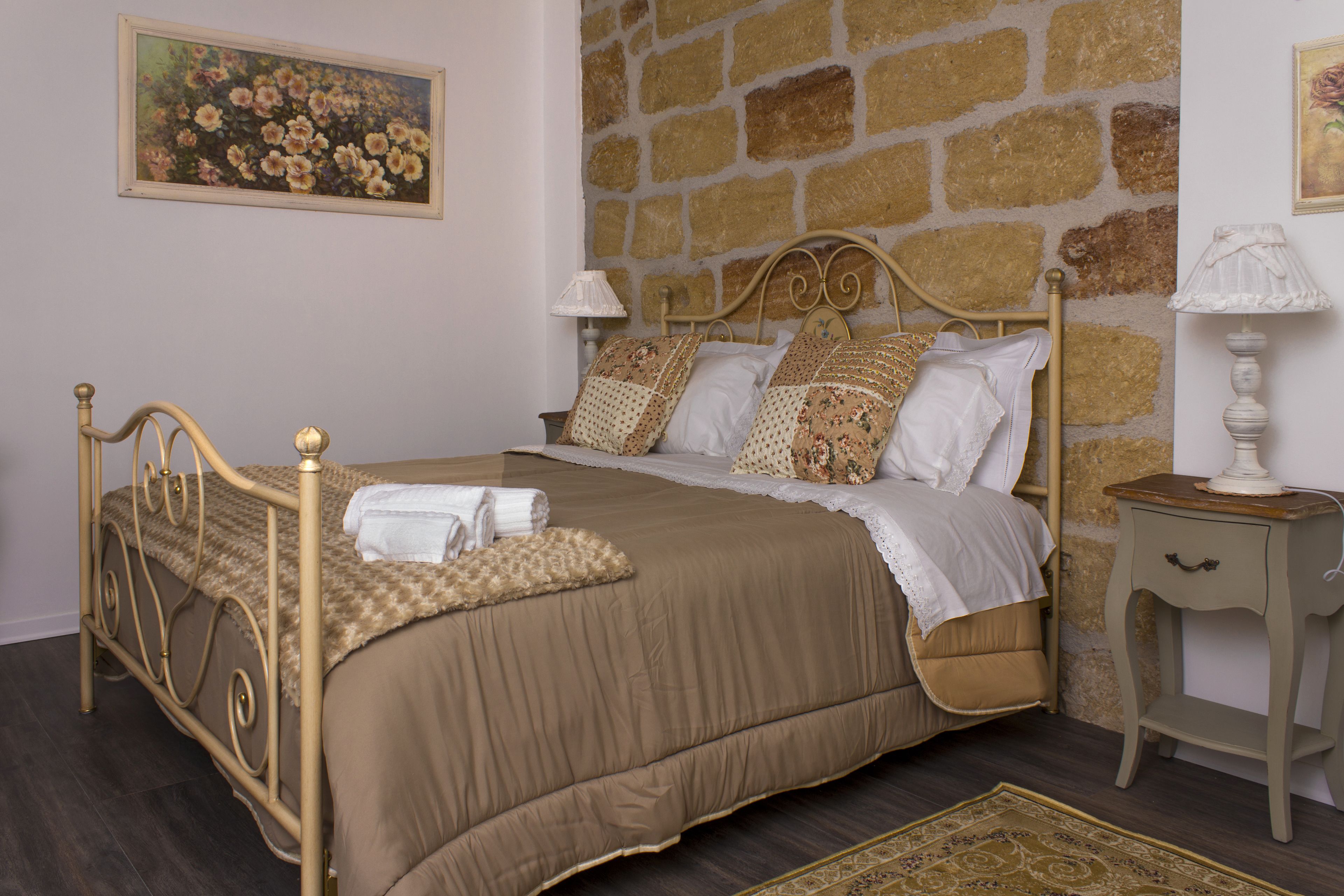 A bedroom with a stone wall, a bed with a beige bedspread, two bedside lamps, and a painting of flowers on the wall.