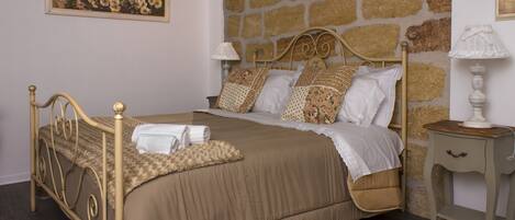 Un dormitorio con pared de piedra, una cama con colcha color crema, dos lámparas de noche y un cuadro de flores en la pared.