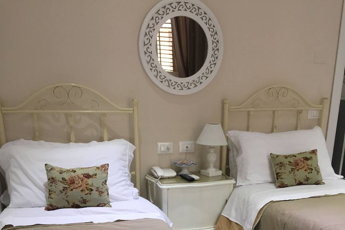 A hotel room with two beds, each with a floral pillow and a white blanket. There is a round mirror on the wall, a bedside table with a lamp, and a telephone.