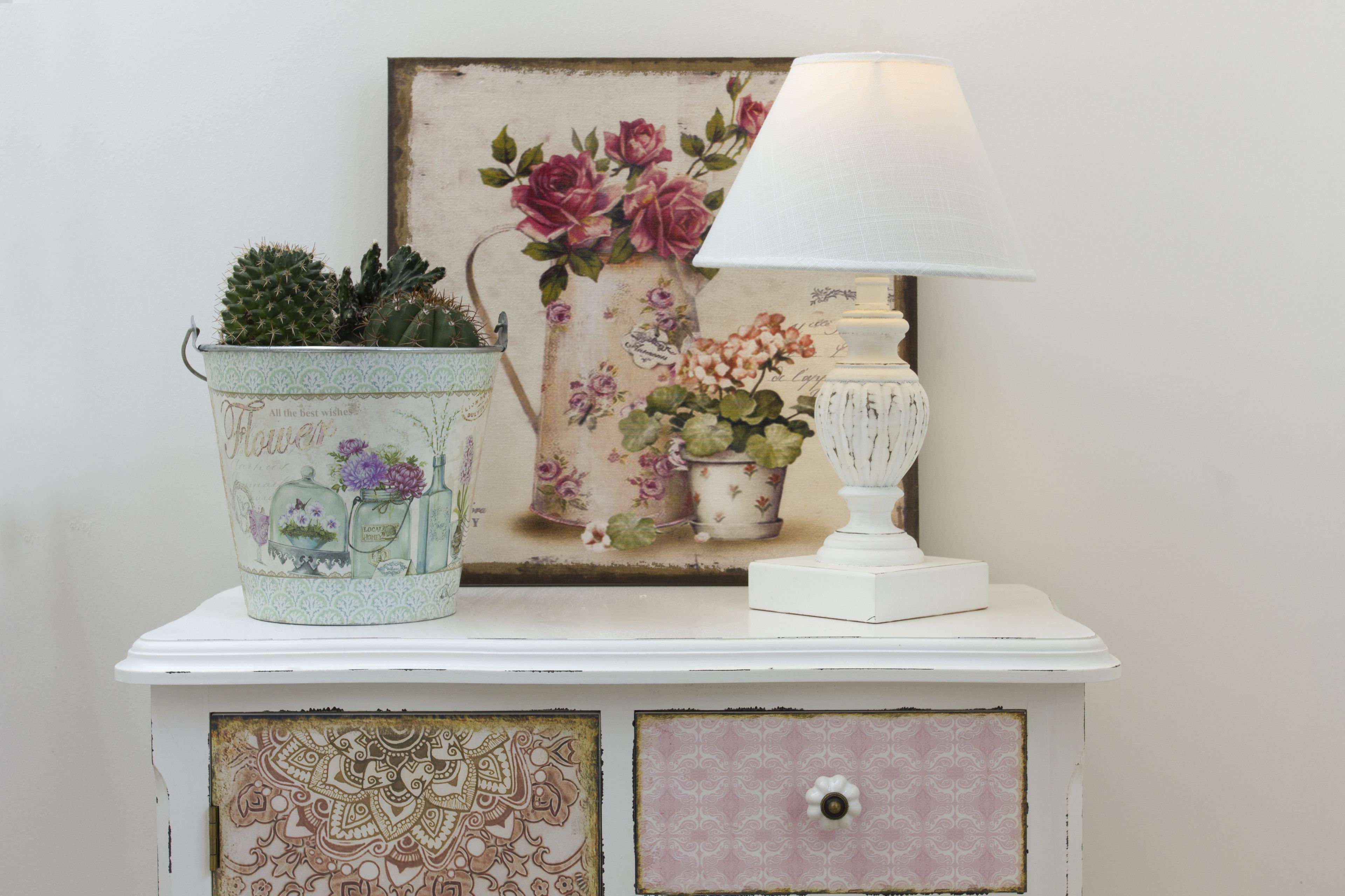 Een witte kast met een bloemschilderij, een lamp in vintage stijl, en een decoratieve pot met cactussen.