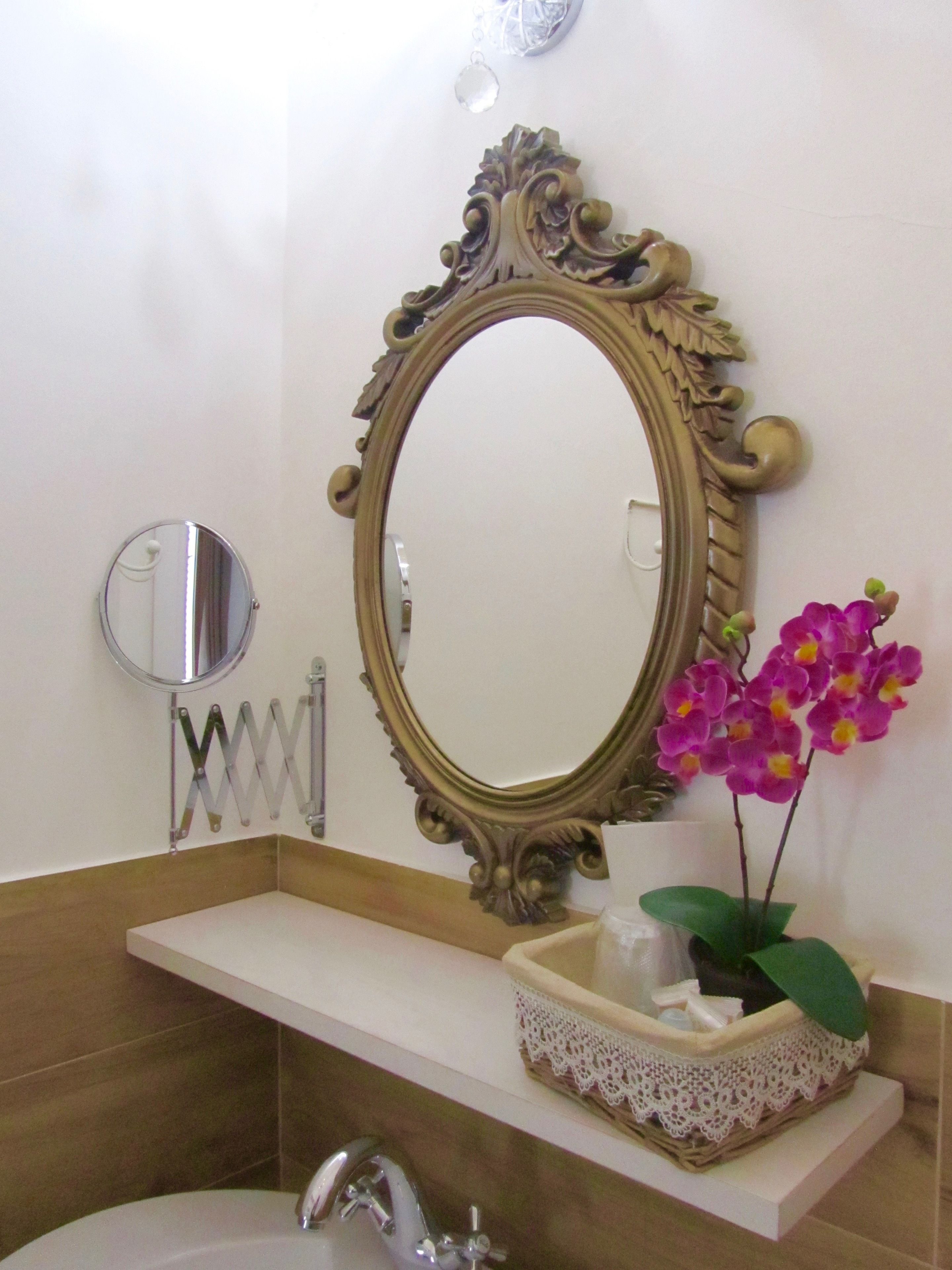 A bathroom with a large ornate mirror, a round mirror, a sink with a lace-trimmed basket, and a potted orchid.