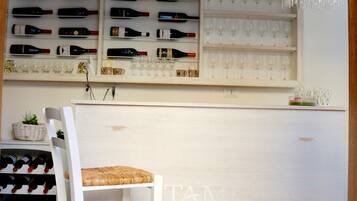 Un espace bar avec des étagères remplies de bouteilles de vin et de verres, un comptoir de bar blanc et une chaise blanche avec un siège en osier tressé.