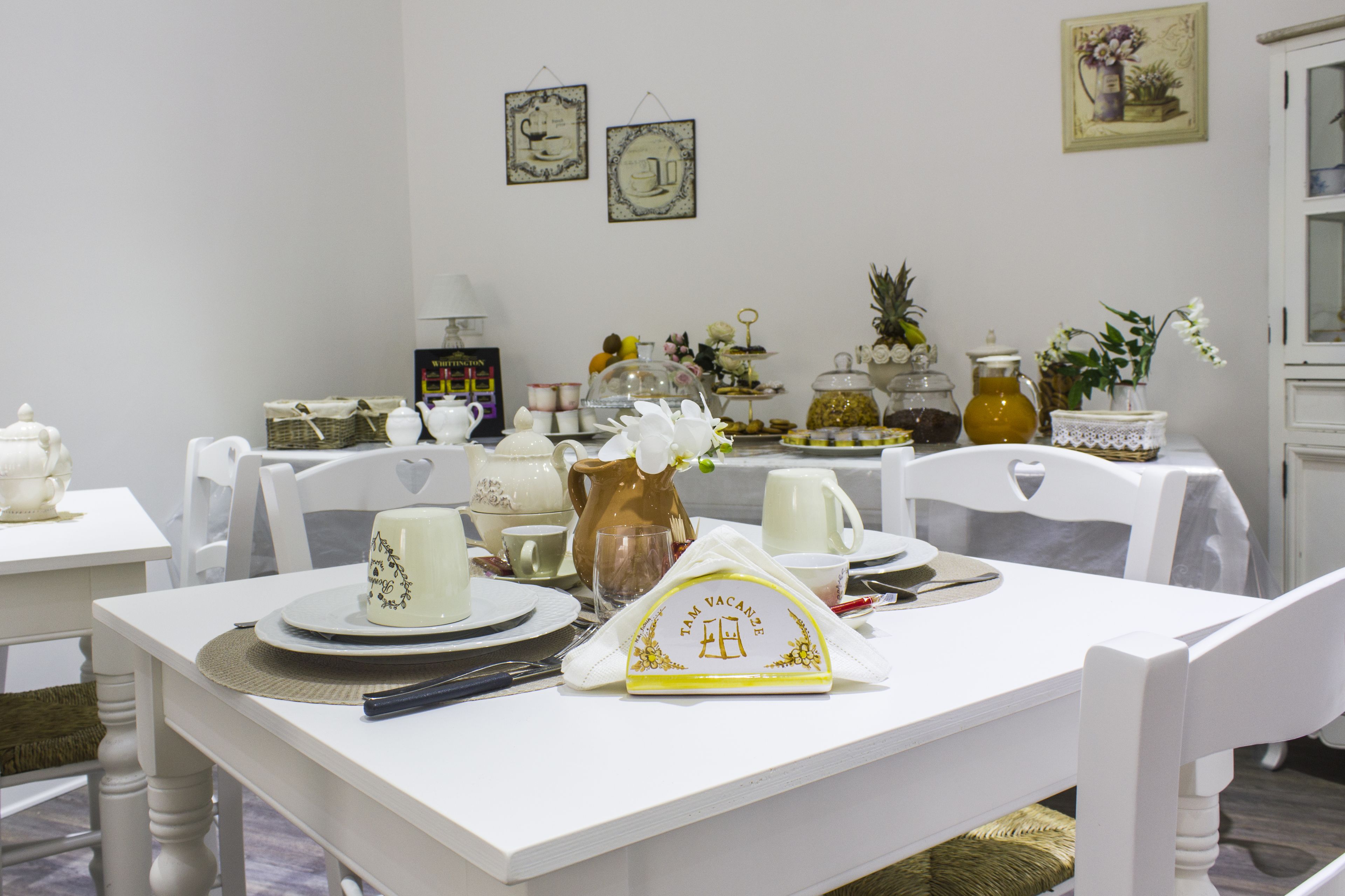 Un comedor con mobiliario blanco, una mesa servida con platos, tazas y una jarra. Se observan cuadros en la pared y un aparador al fondo.