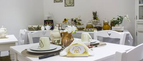 Un comedor con mobiliario blanco, una mesa servida con platos, tazas y una jarra. Se observan cuadros en la pared y un aparador al fondo.