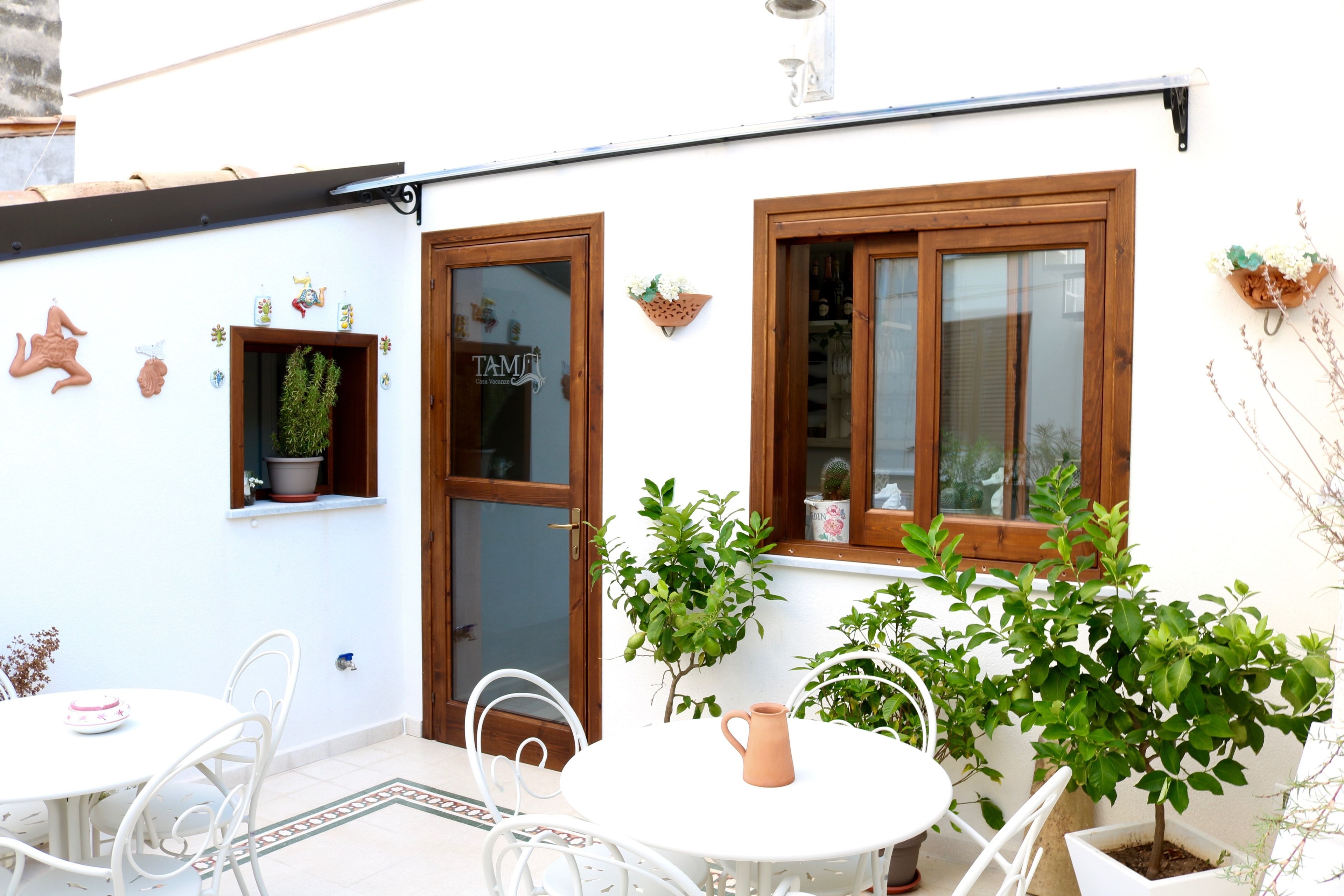 A white building with a wooden door and windows, a small table with chairs, and potted plants.