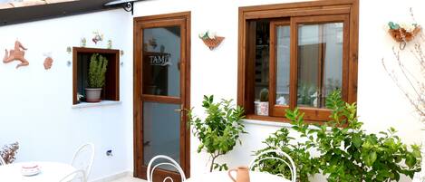 A white building with a wooden door and windows, a small table with chairs, and potted plants.