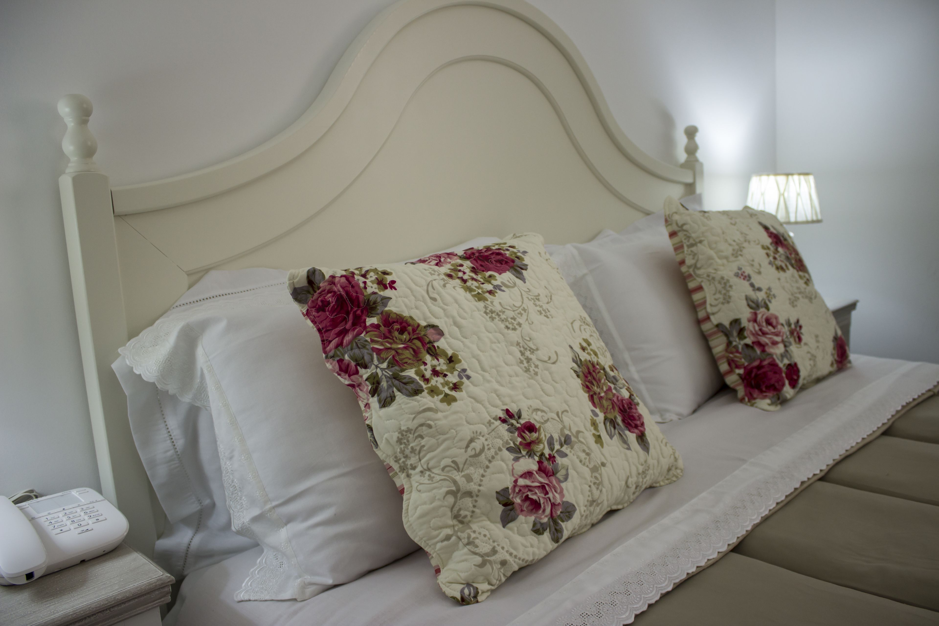 A bed with a floral-patterned quilt and pillows, a bedside table with a phone, and a lamp on the wall.