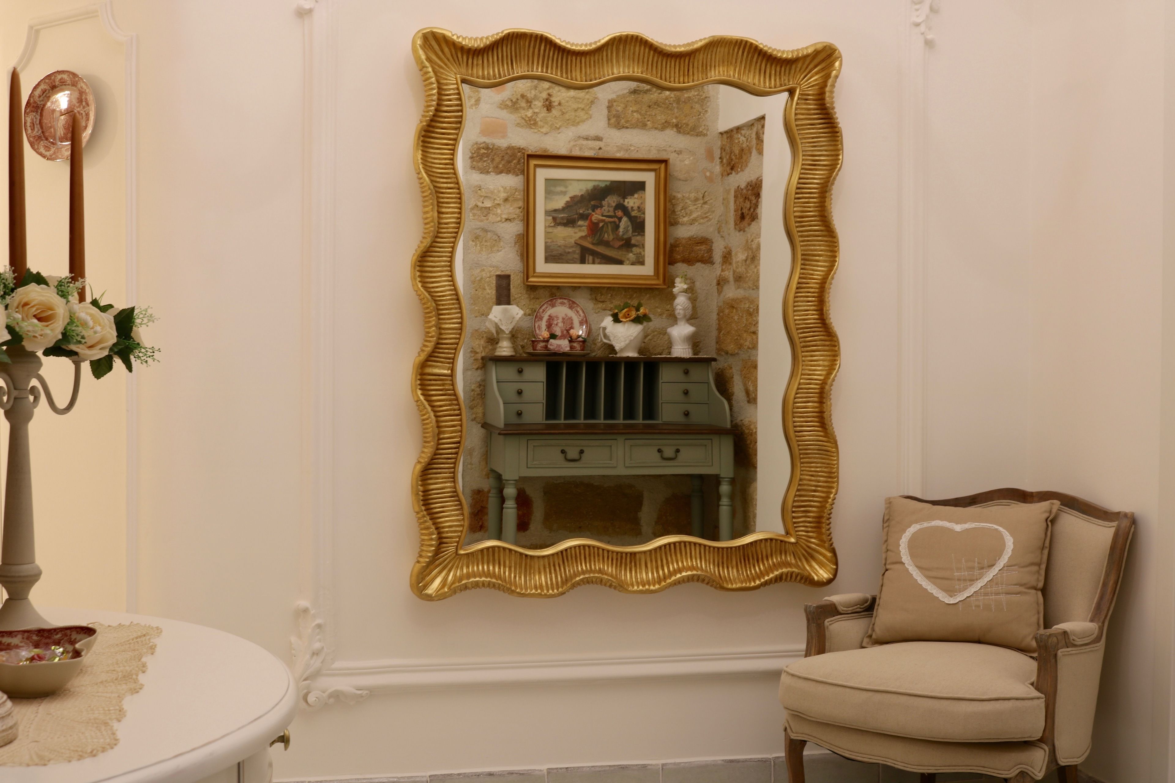 A room with a large ornate mirror, a small vanity with drawers, a chair with a heart-shaped pillow, and a vase with flowers on a table.