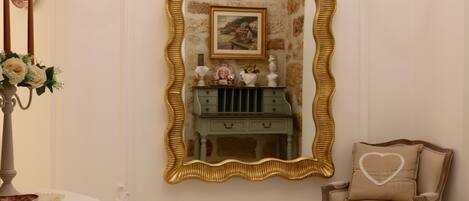 A room with a large ornate mirror, a small vanity with drawers, a chair with a heart-shaped pillow, and a vase with flowers on a table.