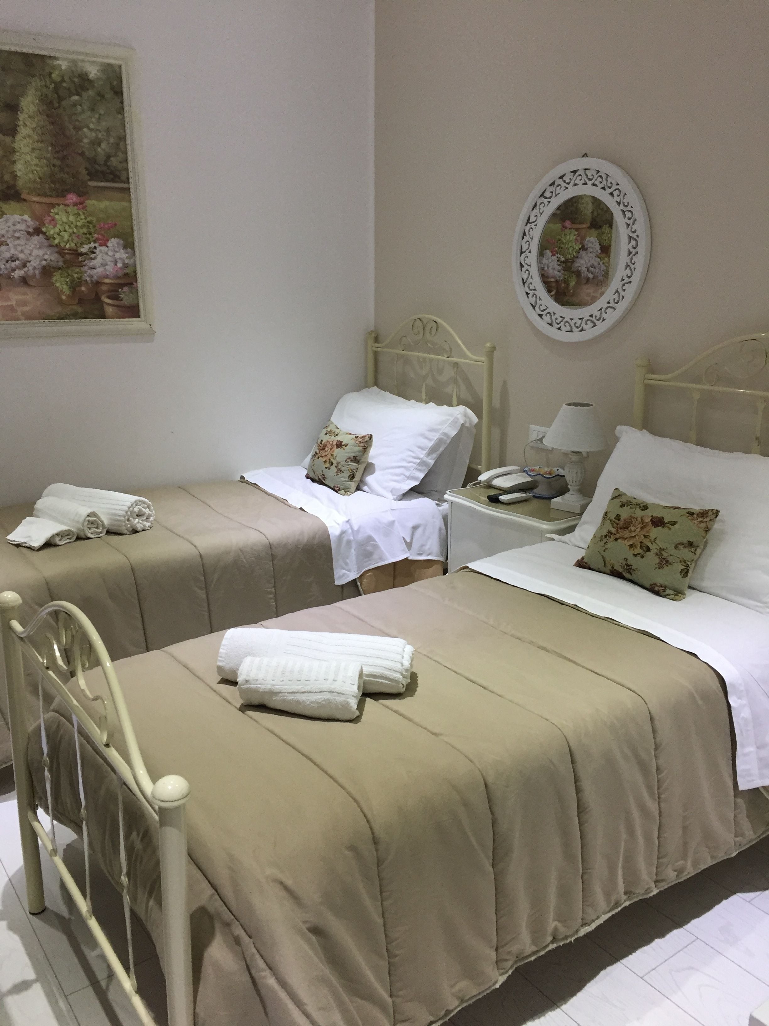 A hotel room with two beds, each with a white pillow and a beige bedspread. There is a small table with a lamp and a telephone.