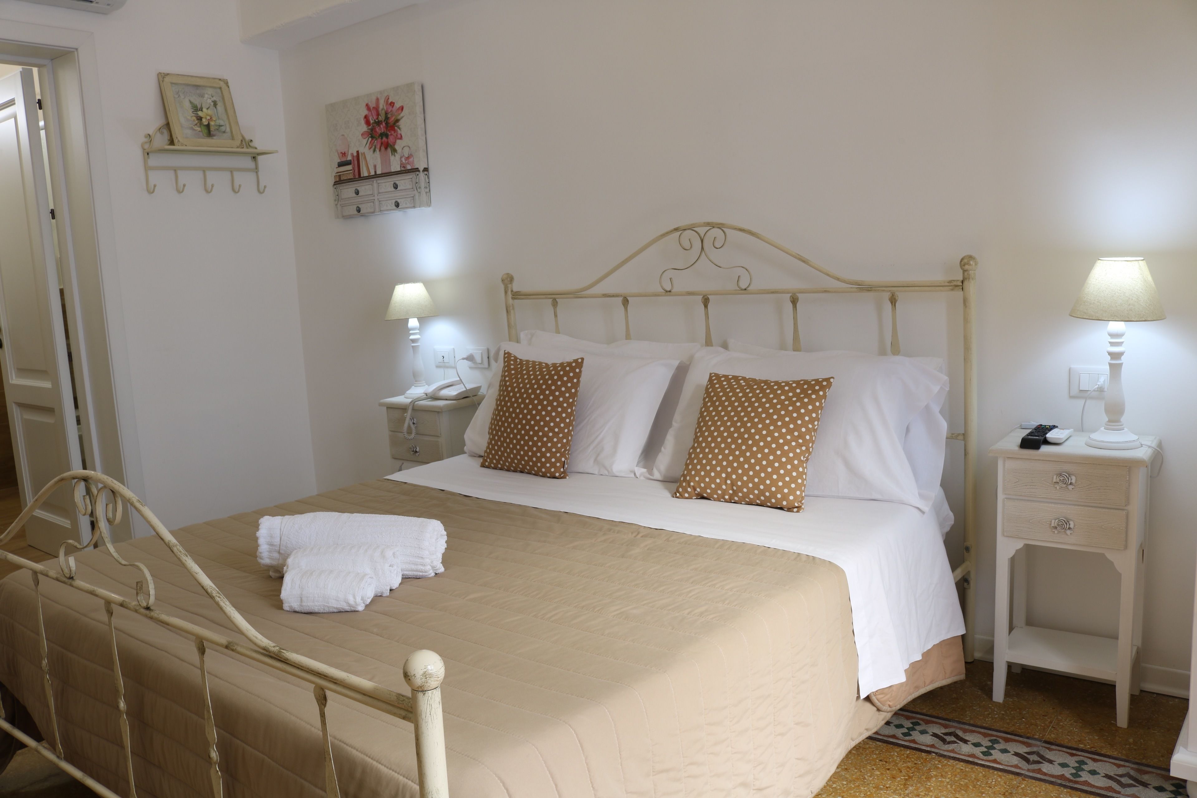 Una cama bien hecha con almohadillas blancas y marrones de lunares, una mesita de noche blanca con una lámpara y un pequeño estante con un cuadro enmarcado.