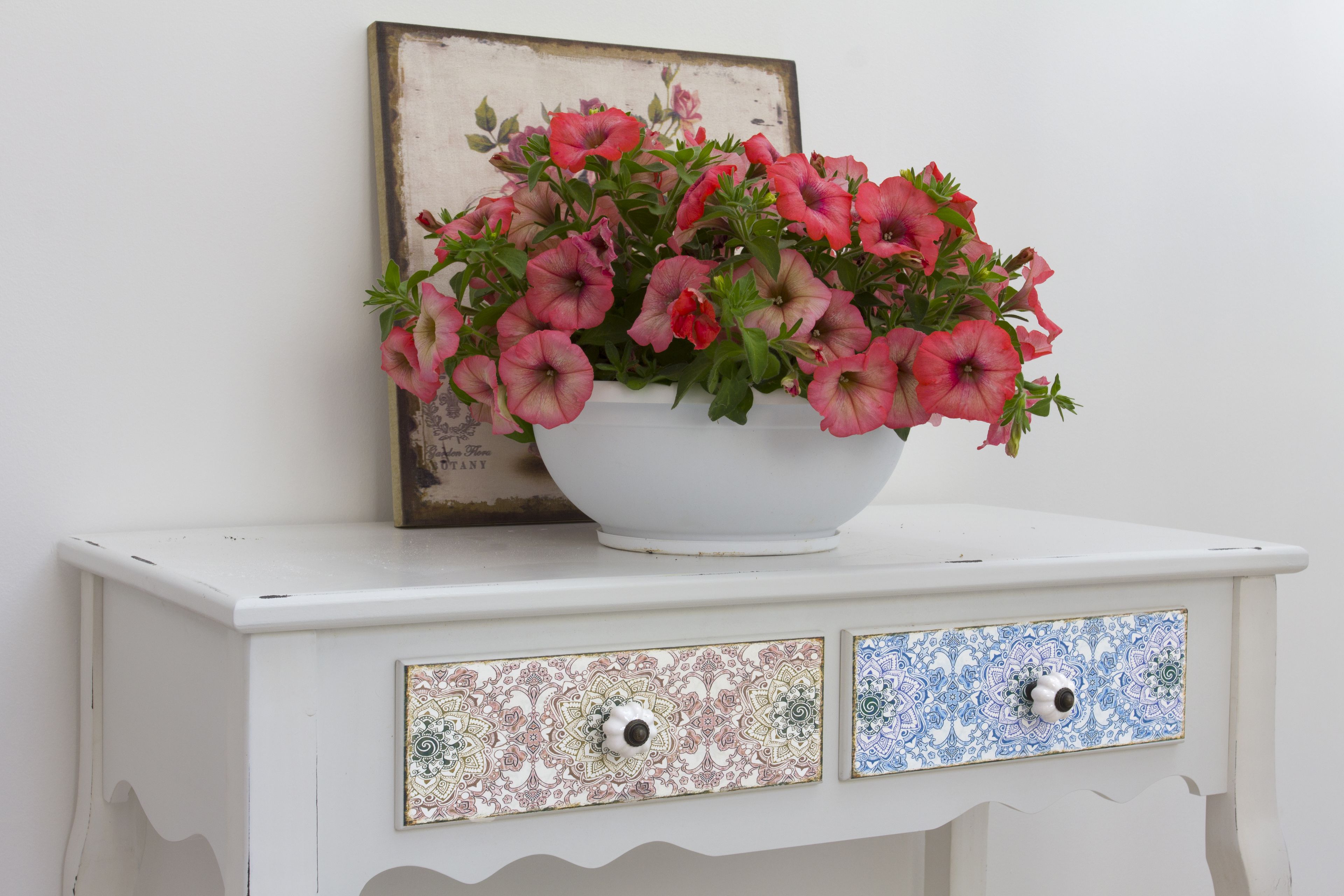 Un aparador blanco con un cuenco blanco que contiene flores rosas, dos cajones con azulejos estampados y un cuadro en la pared.