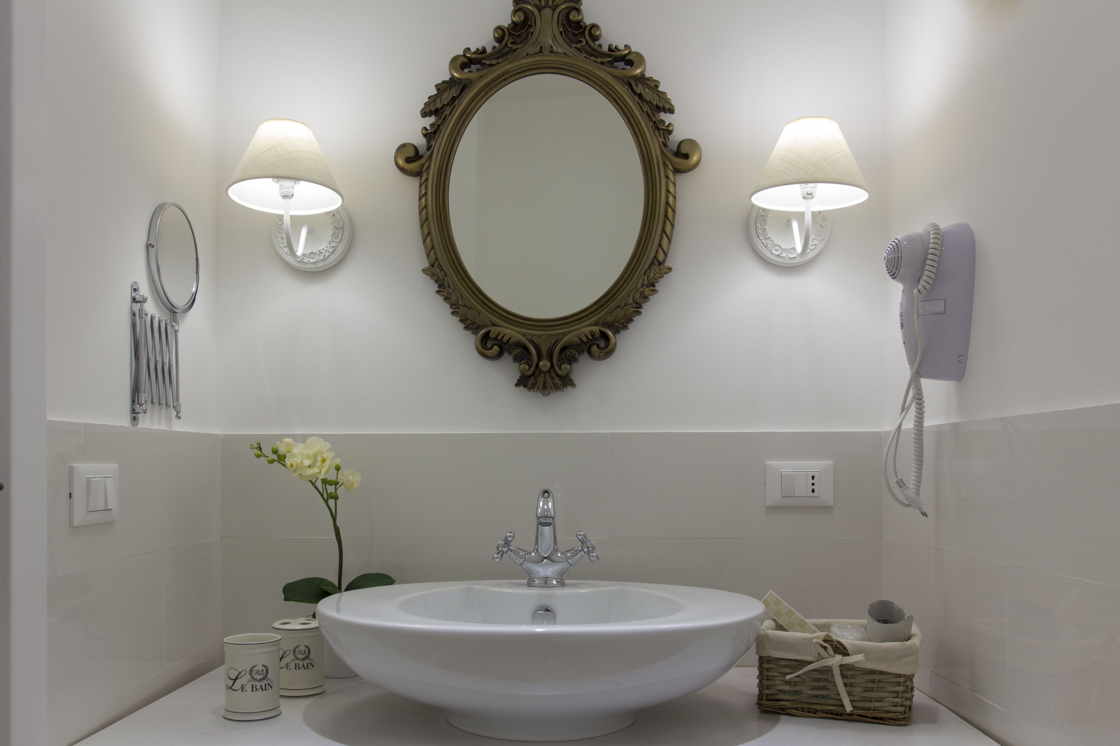 A bathroom with a large oval mirror, two wall-mounted lamps, a white sink, a hairdryer, and a small wicker basket.